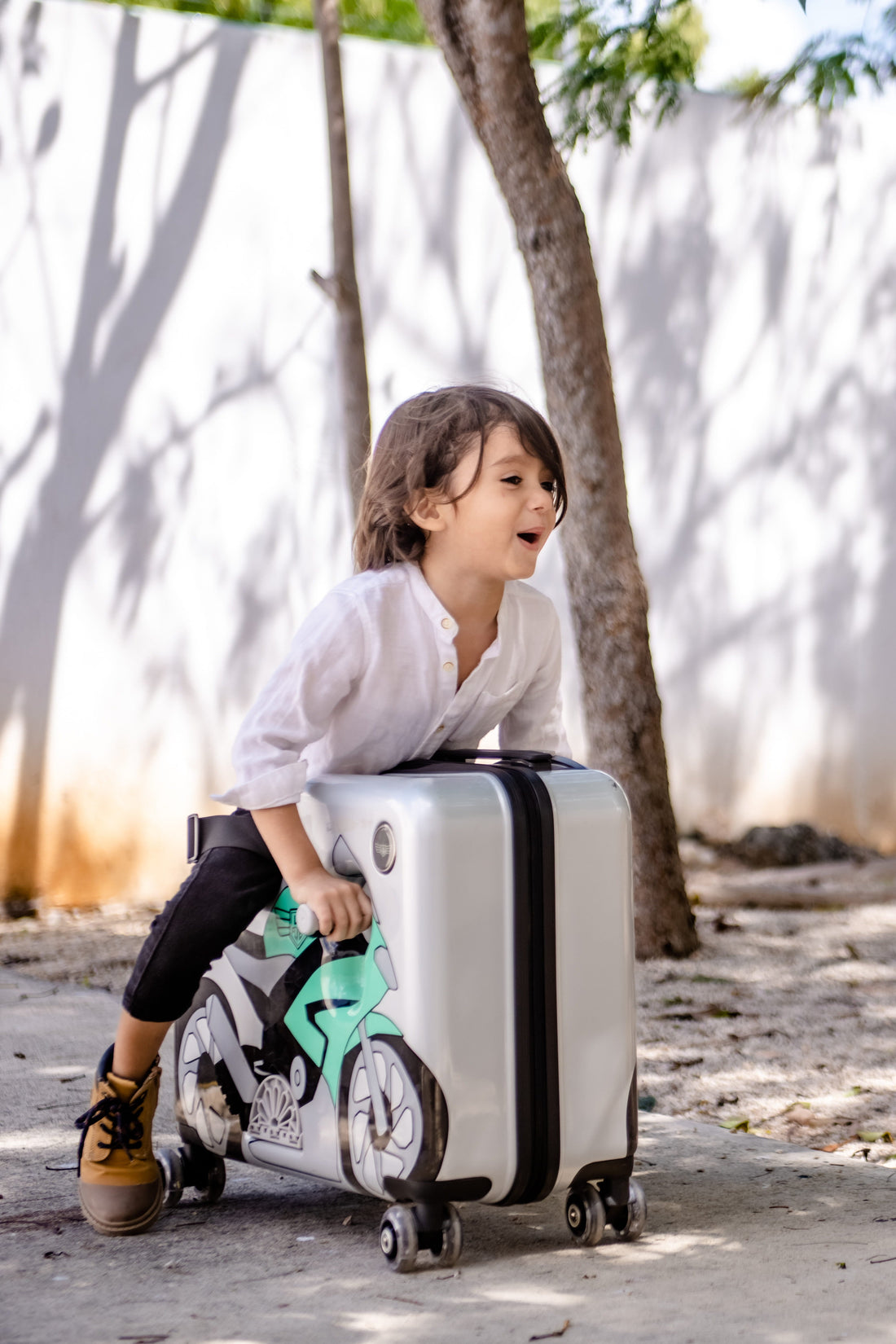 A child leaning over a white ride-on suitcase with a motorcycle design near trees and a white wall outdoors.