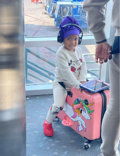 A young girl wearing black headphones, a blue bonnet, and a gray sweatsuit sits on a suitcase stroller at an airport.
