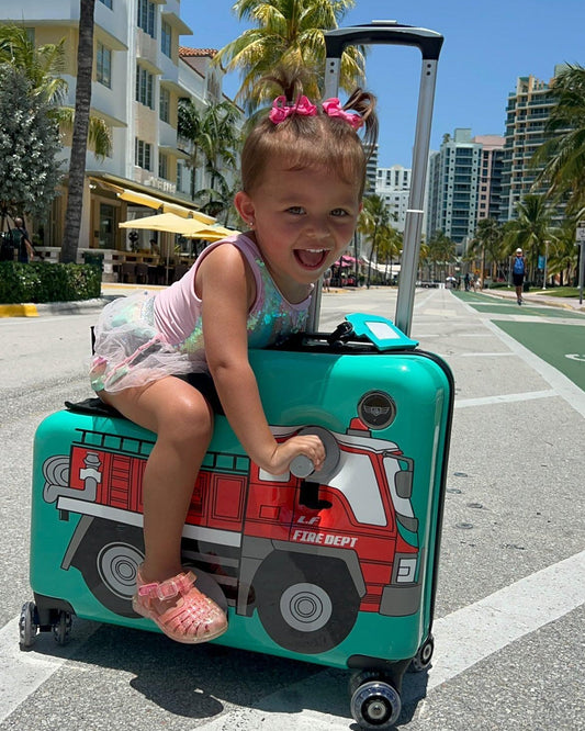 Child sitting on a teal ride-on suitcase with a fire truck graphic on a sunny city street lined with palm trees.