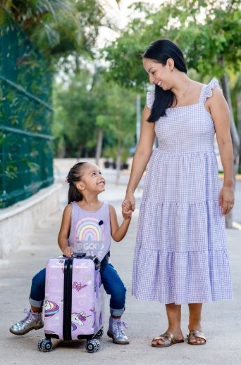 A little girl smiles up at her mom as she holds her hand and sits on a purple suitcase. Mom is standing next to her.