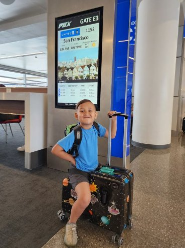 A preschool-aged boy wearing a blue shirt and gray shorts sits on a space-themed ride-on suitcase next to an airport gate.