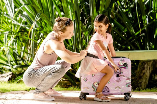A little girl sits on a ride-on suitcase that is pink and has unicorns. She is looking over her shoulder at her mom.
