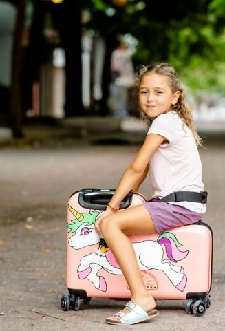 An elementary school-aged girl sits on a pink ride-on luggage that has a white unicorn on it and a rainbow tail.