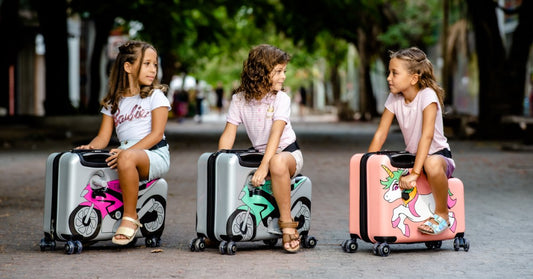 Three children sit on ride-on suitcases decorated with colorful graphics, facing one another on a tree-lined street.