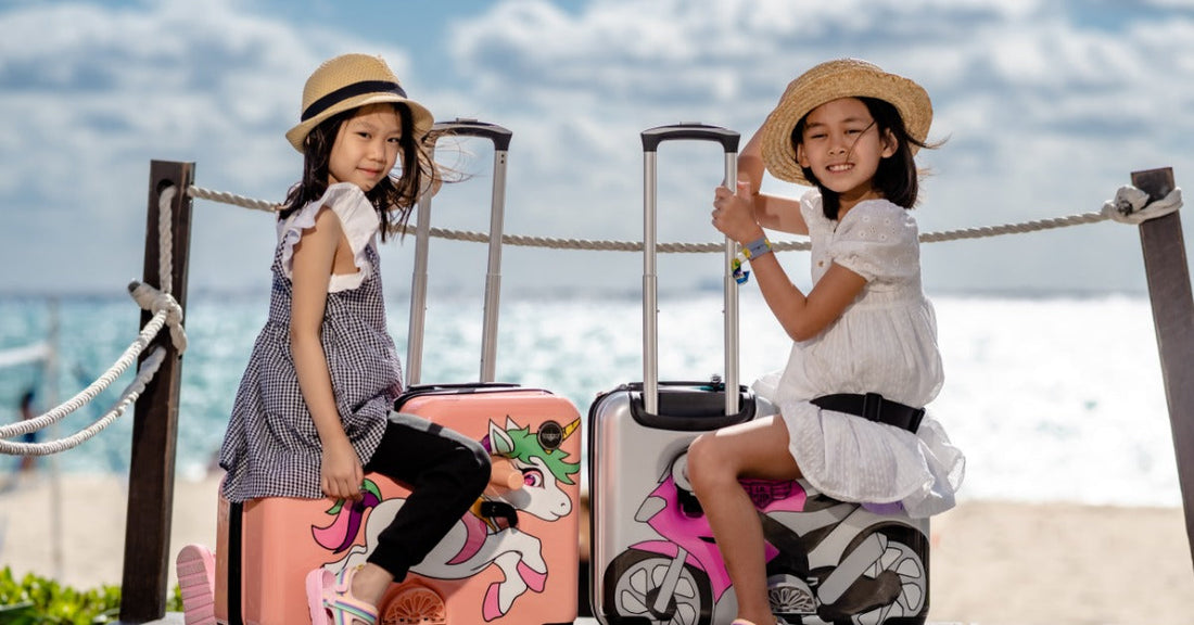 Two children wearing straw hats sitting on colorful ride-on suitcases near a rope fence by an ocean beach.